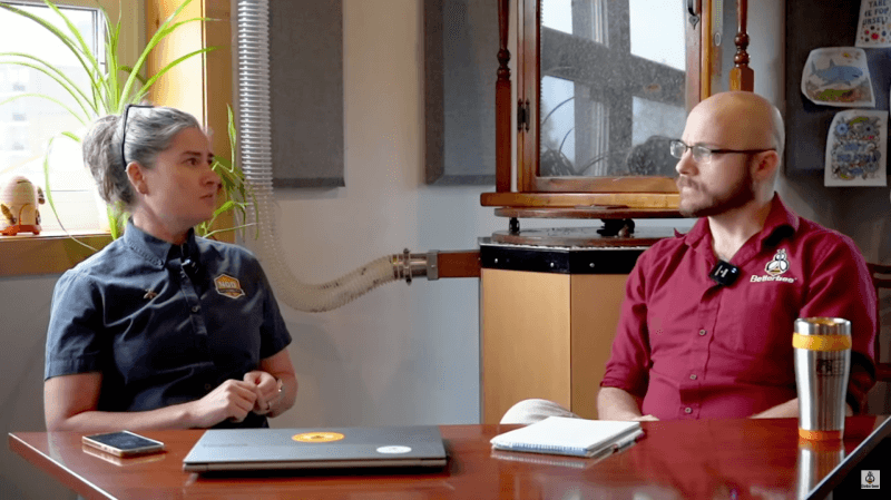 Two people sit across a table discussing beekeeping topics in an interview setting, with notebooks, a laptop, and a travel mug on the table.